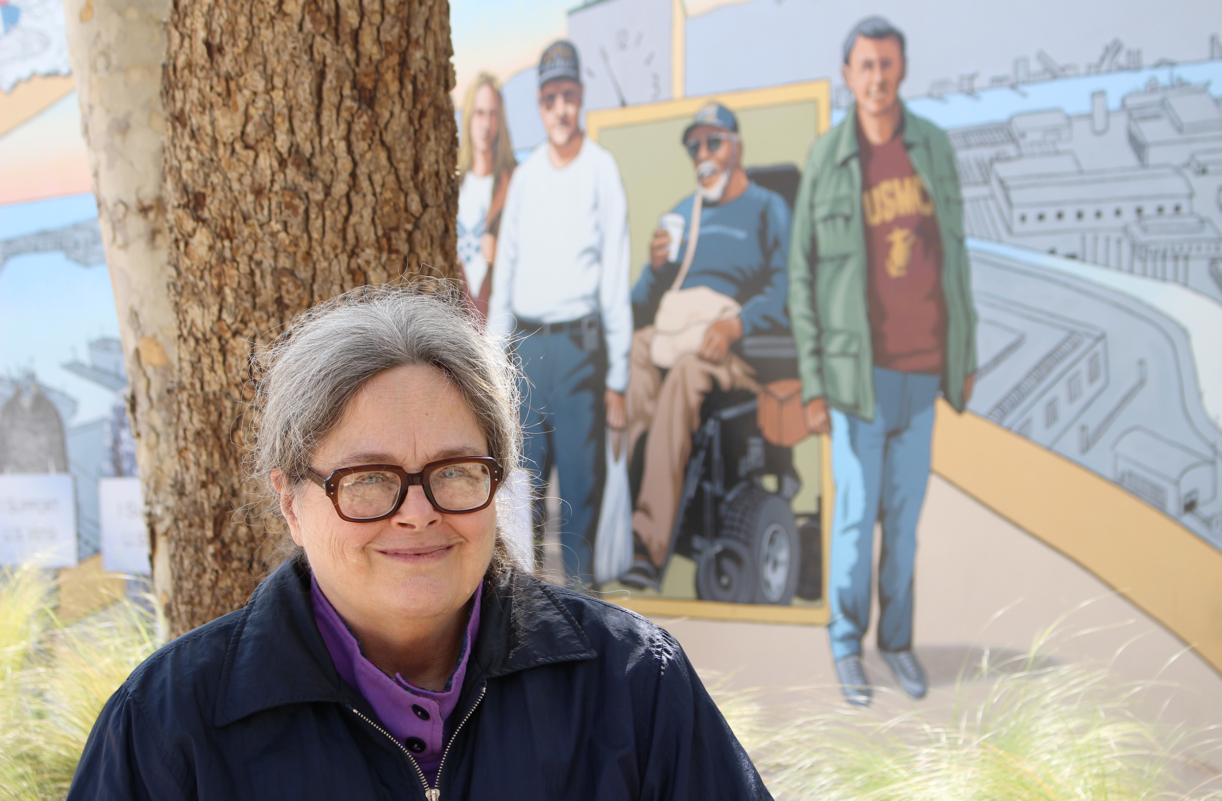 A woman sits in front of a tree and a mural of several people.
