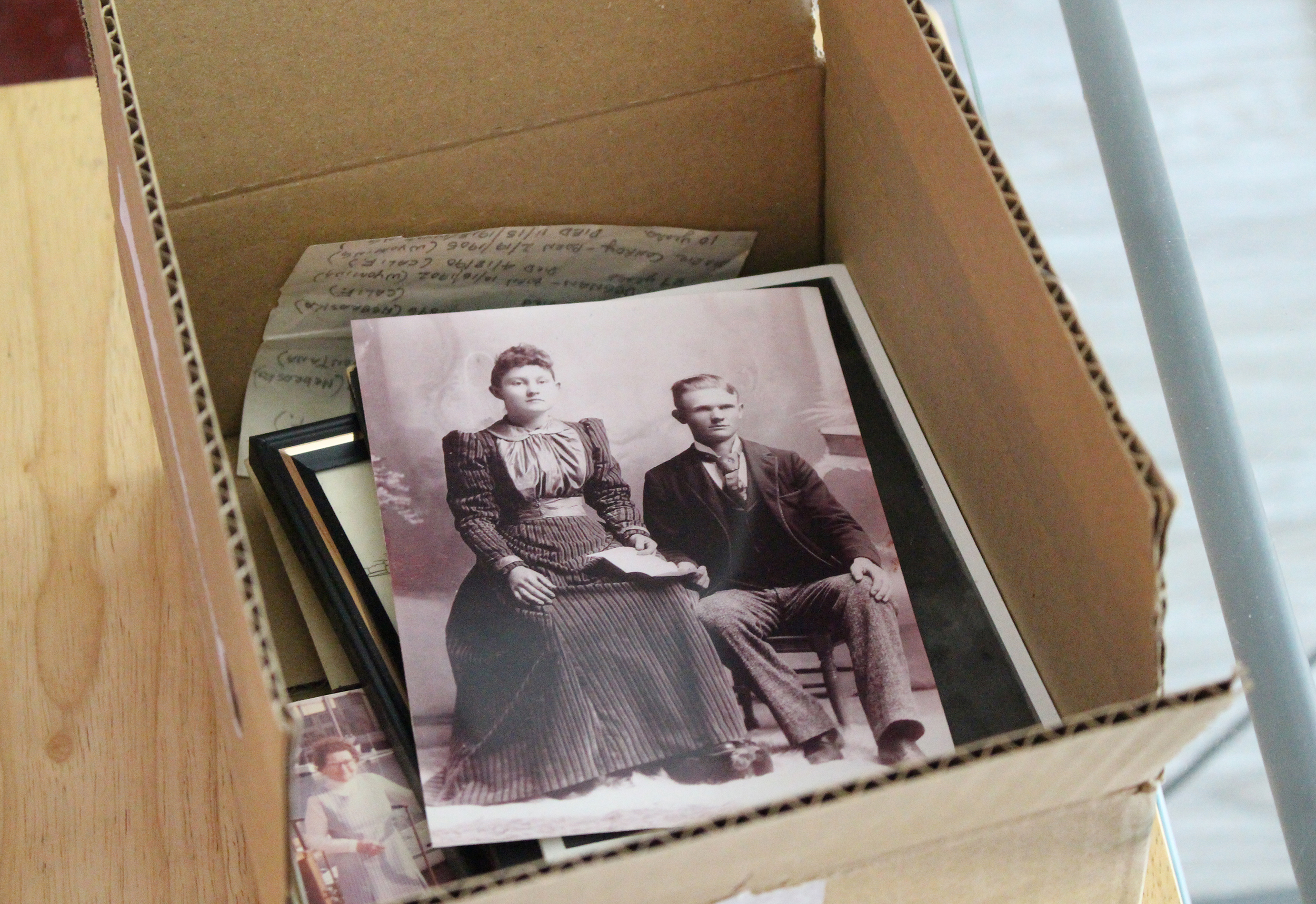 A cardboard box holds several photos and notes. A picture of a man and a woman posing for a portrait is shown at the top.