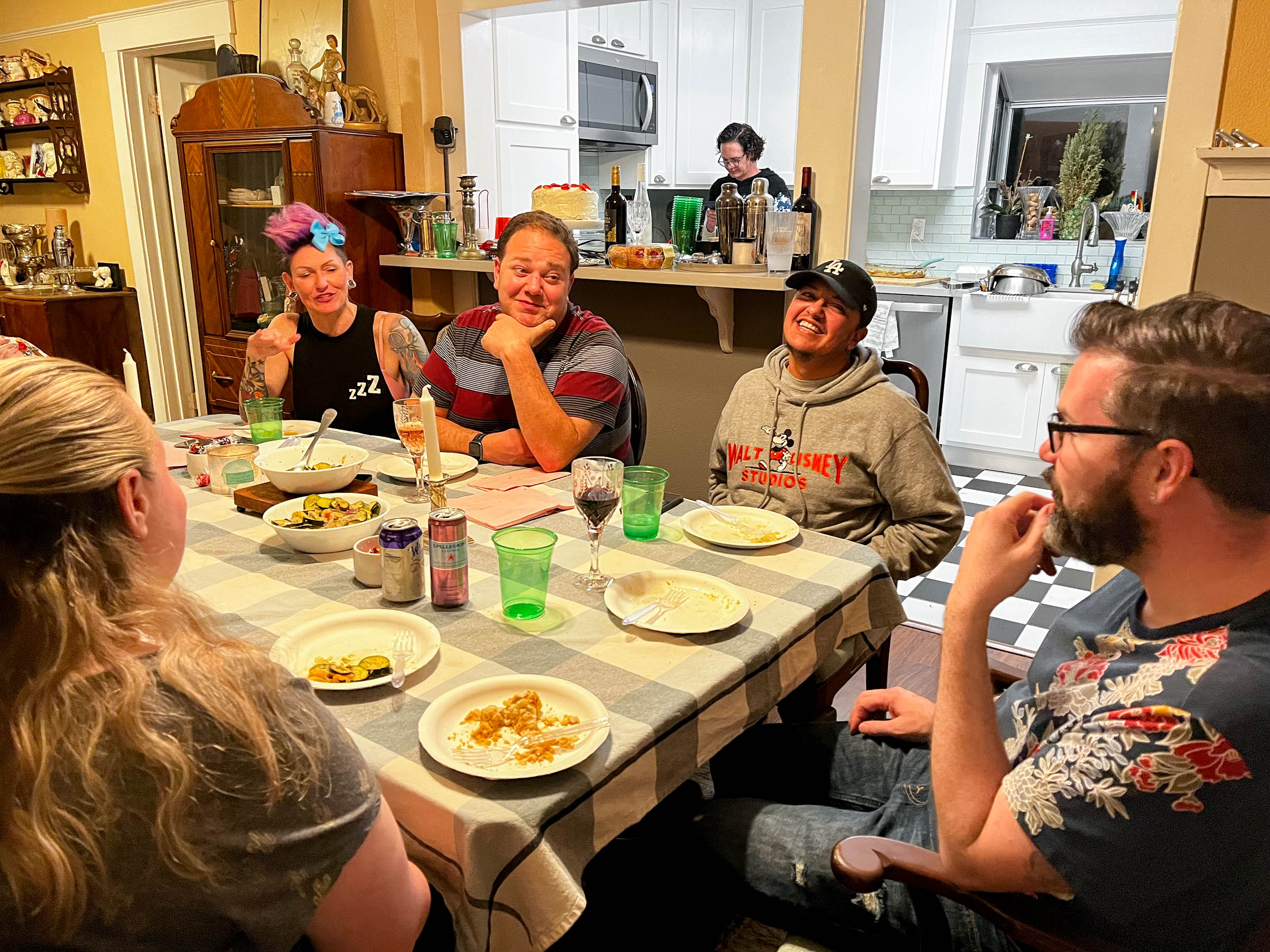 Five people sit around a dining room table smiling at eachother with plates of food in front of them. One can be seen through a window into the kitchen.