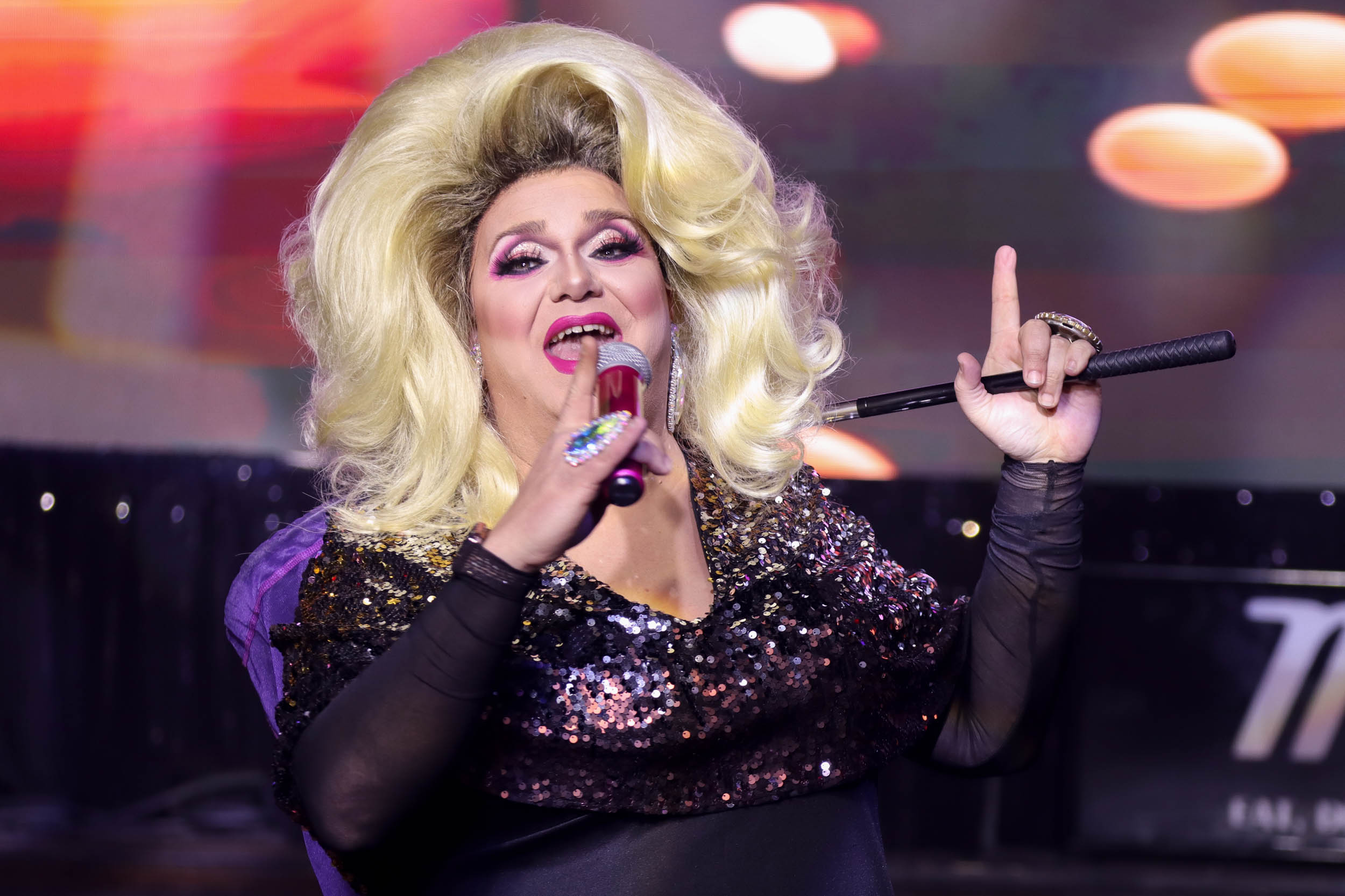 A drag queen in a large blonde wig speaking into a microphone as she addresses the crowd. In her other hand she holds a purple butterfly net behind her head used to collect money.