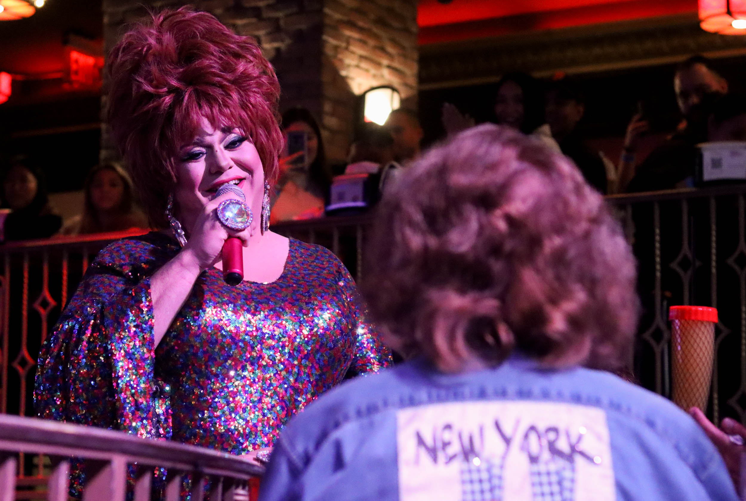 A red headed drag queen in the spotlight dressed in a sequined, colorful top sings to customer. A person sits in the foreground with her back to the camera, with curly hair and a denim jacket with a ‘New York’ patch on the back.
