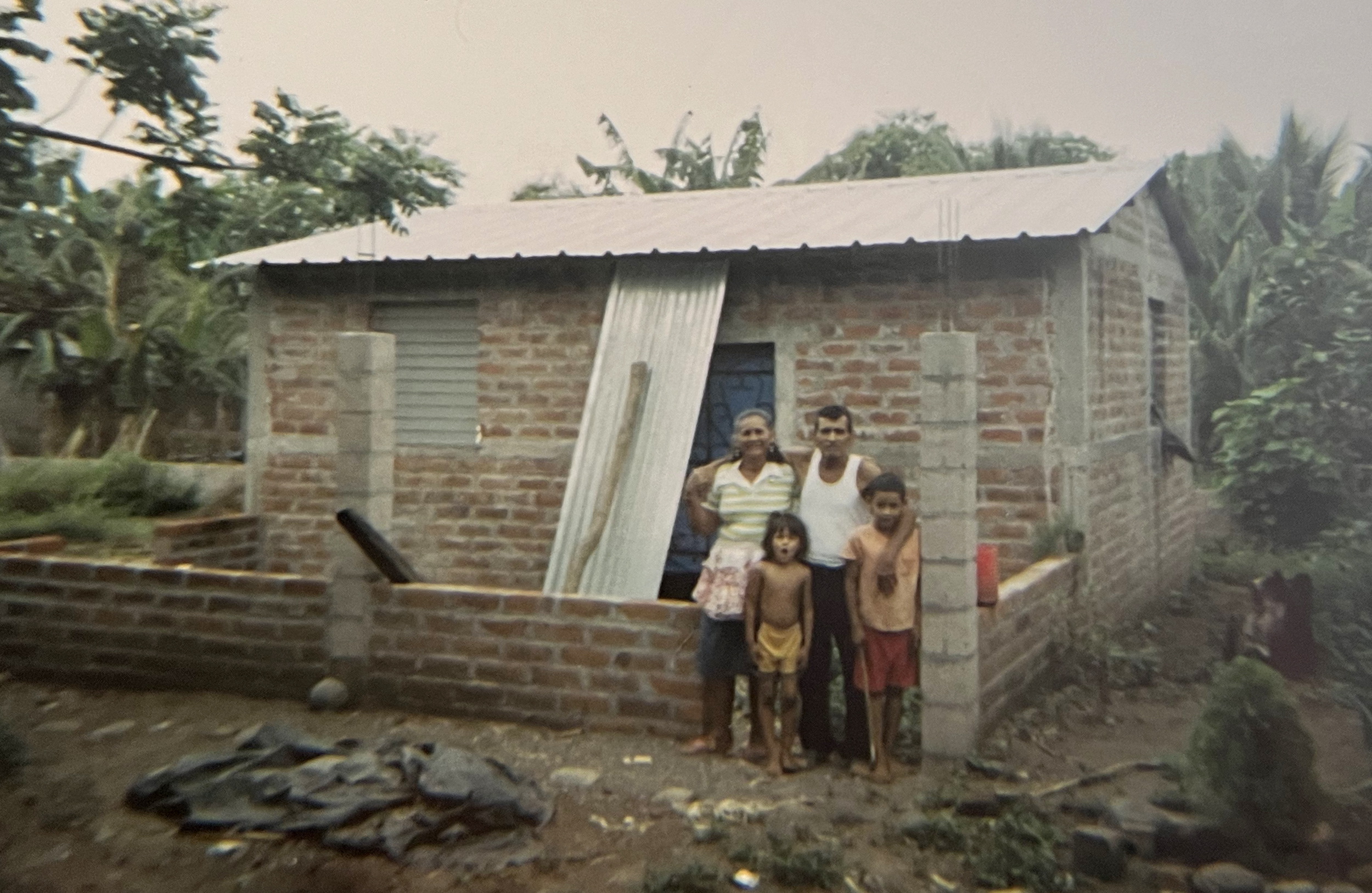Two adults stand behind and embrace two children as they all pose in front of a brick house surrounded by trees.