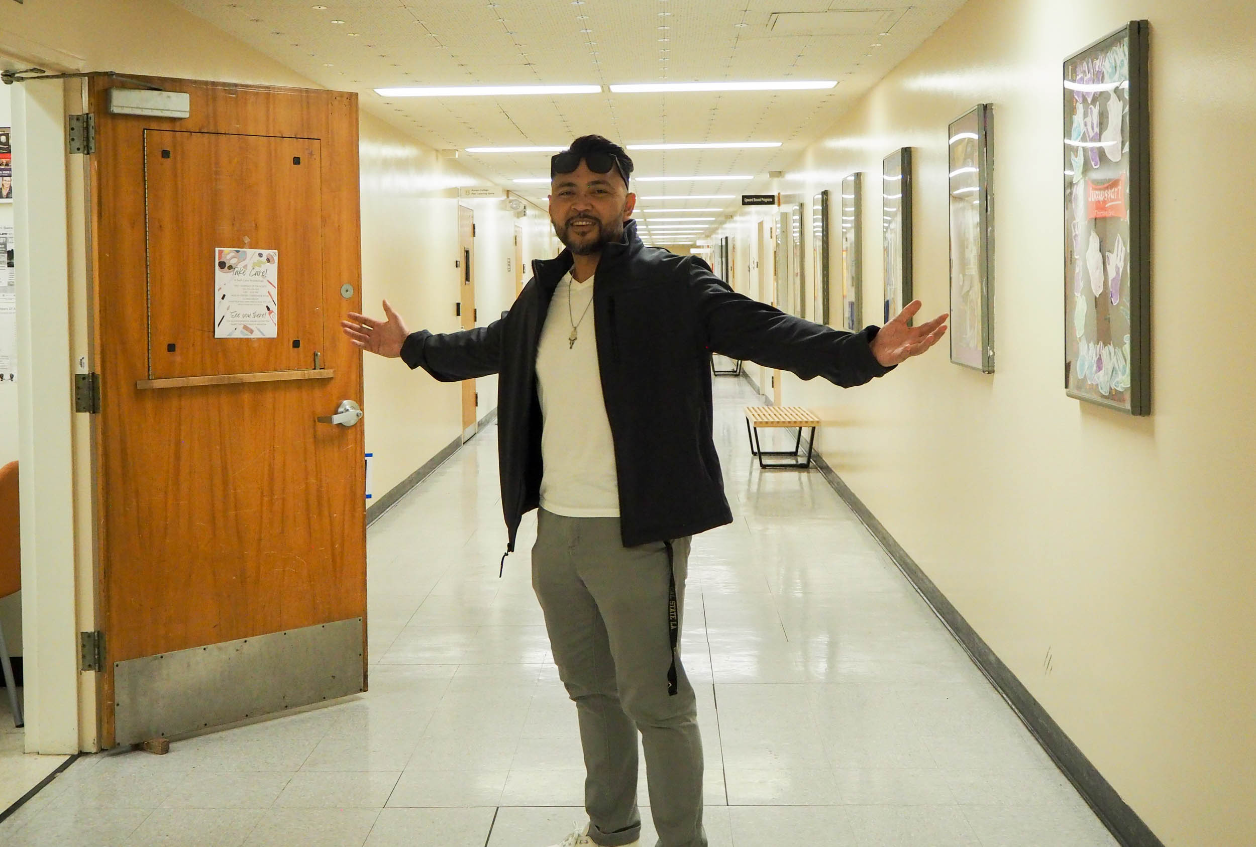 A man stands in the middle of a long university hallway with open arms.