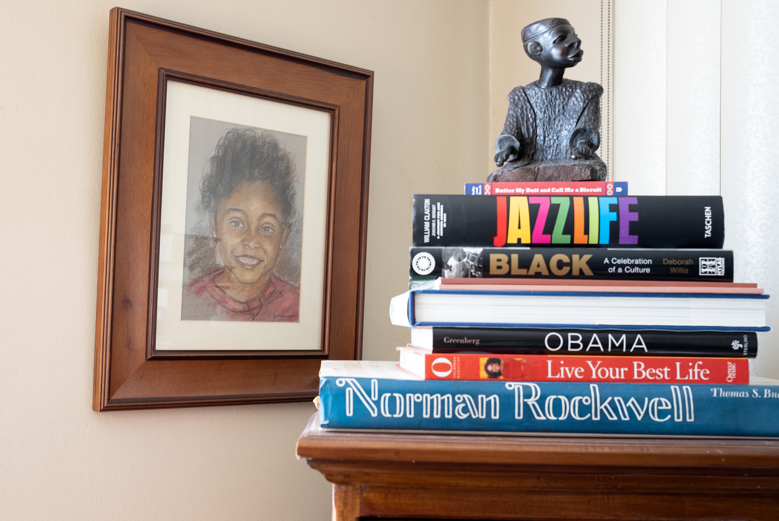 A drawing of a young girl hangs on the wall next to a stack of books and a sculpture on top of a cabinet.