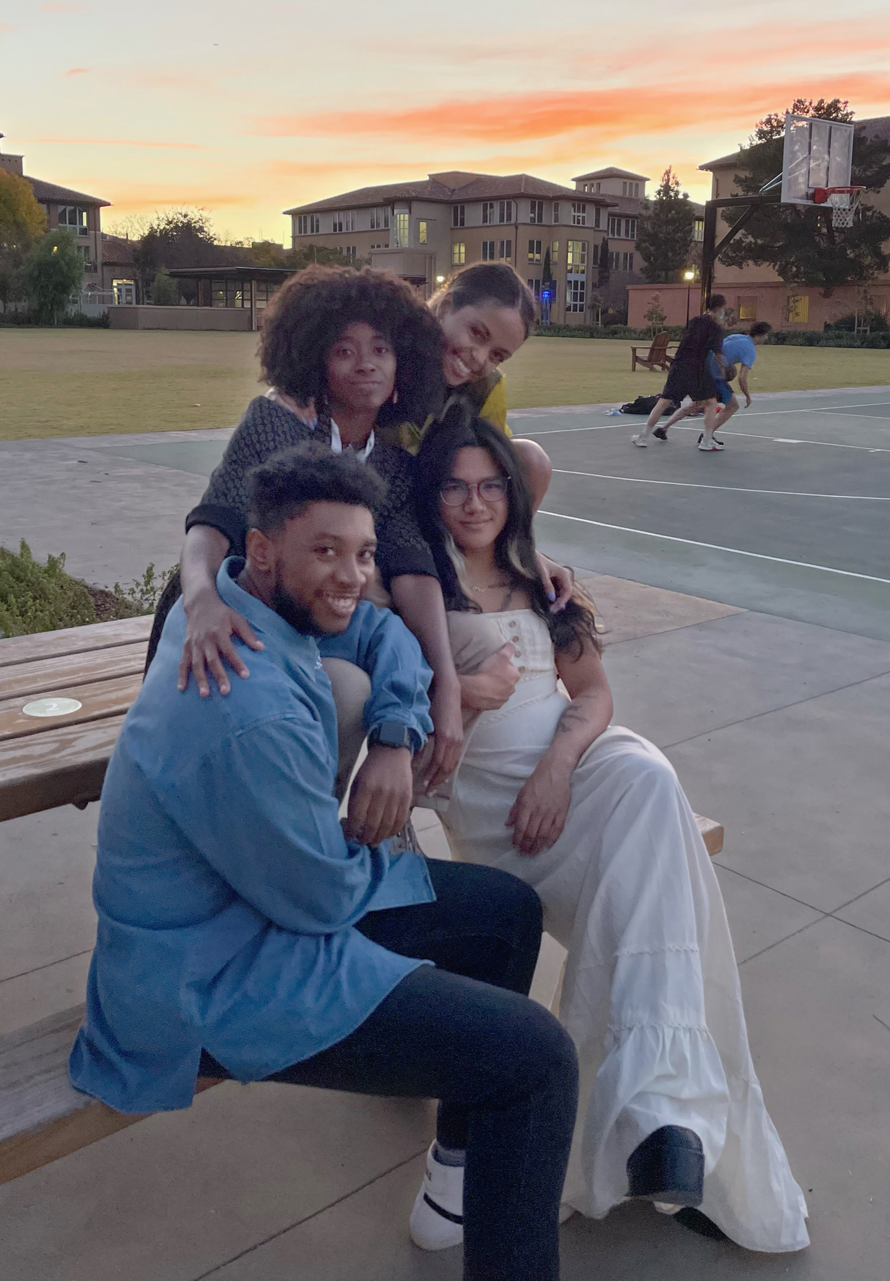 A group of four friends sit on a table in a basketball court, embracing each other. Behind them, the sunset creates orange clouds in contrast to the light blue sky.